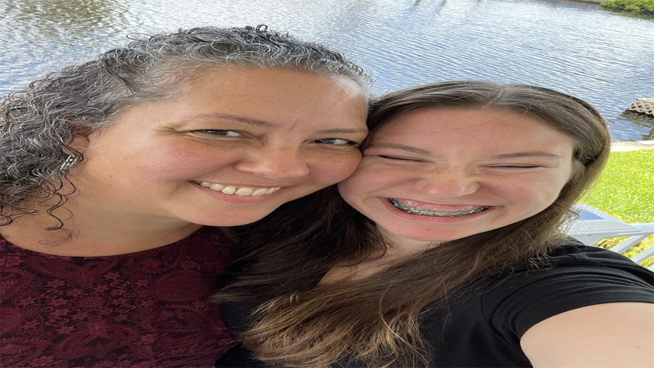 Giselle Espinet (left) and her adopted daughter, Liliana, hug each other tightly, cheek to cheek. Liliana’s arm is raised to take the selfie-style portrait of the two.