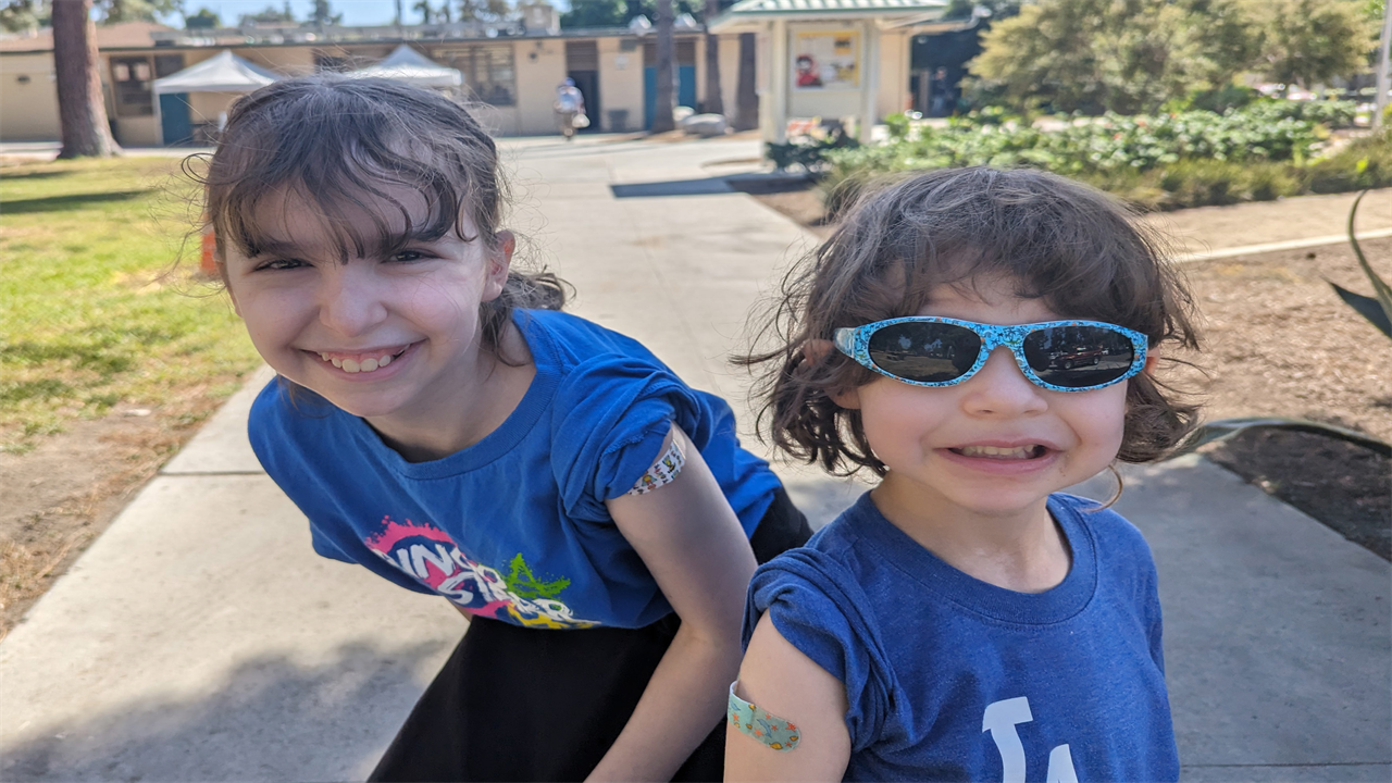 A photo of two children smiling for the camera showcasing bandages on their arms after getting vaccinated.