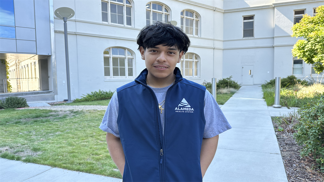 A young man wearing a vest with the words "Alameda Health System" on it stands outside of a building and looks at the camera.
