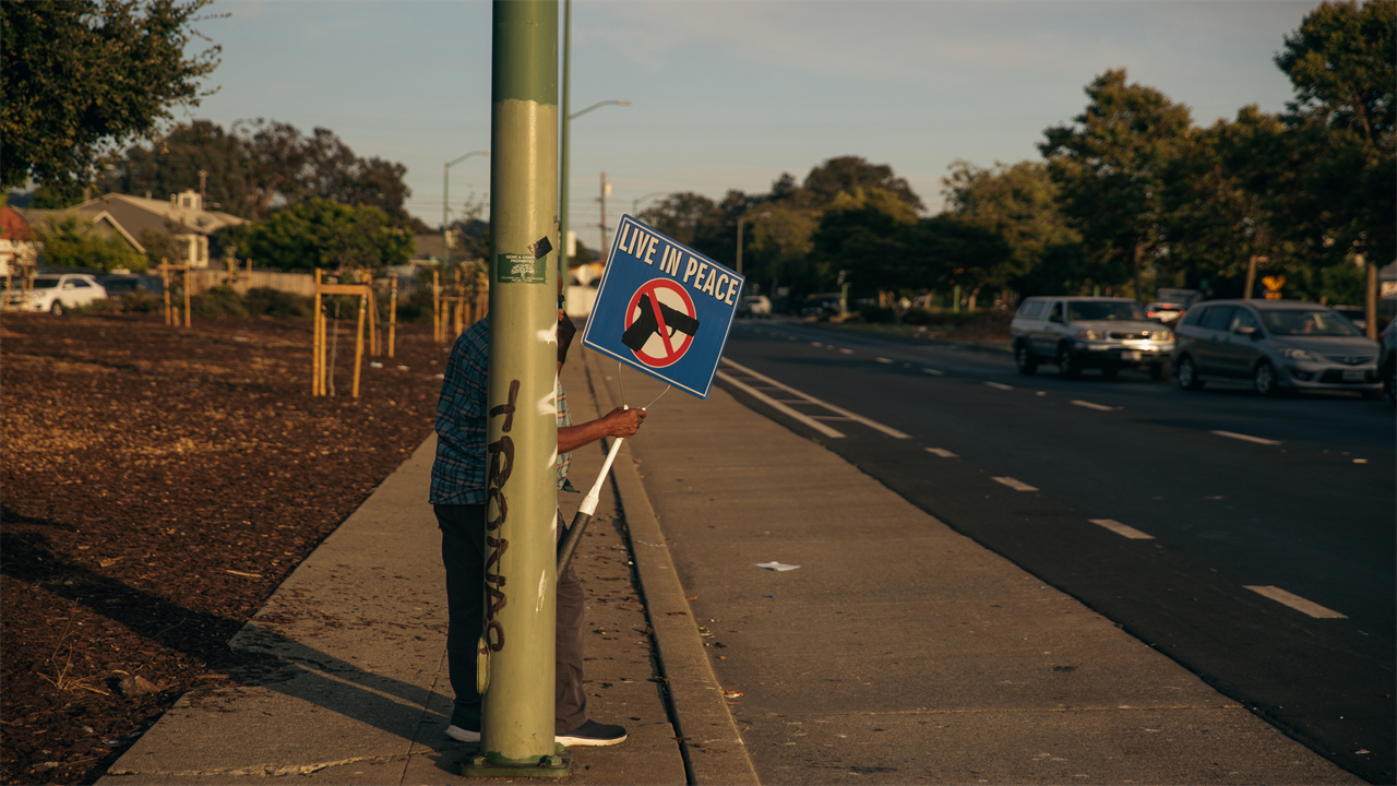 A person holds a sign that reads "Live In Piece" and has the image of a handgun behind a general prohibition sign. They stand behind a streetlight pole beside a road.
