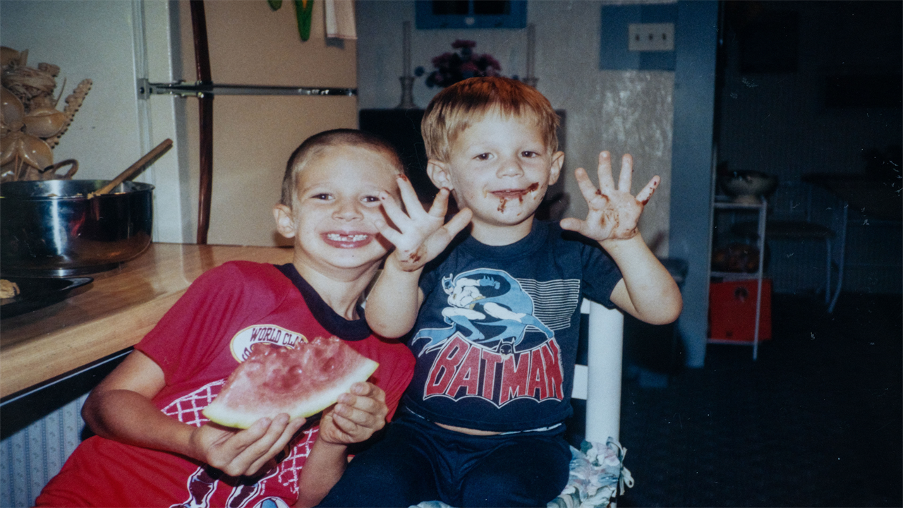 Two children, both boys, smile at the camera. One boy, in a red tshirt, is eating a slice of watermelon and has several teeth missing. The other, in a navy Batman tshirt, holds up his hands which, like his face, are covered in chocolate.