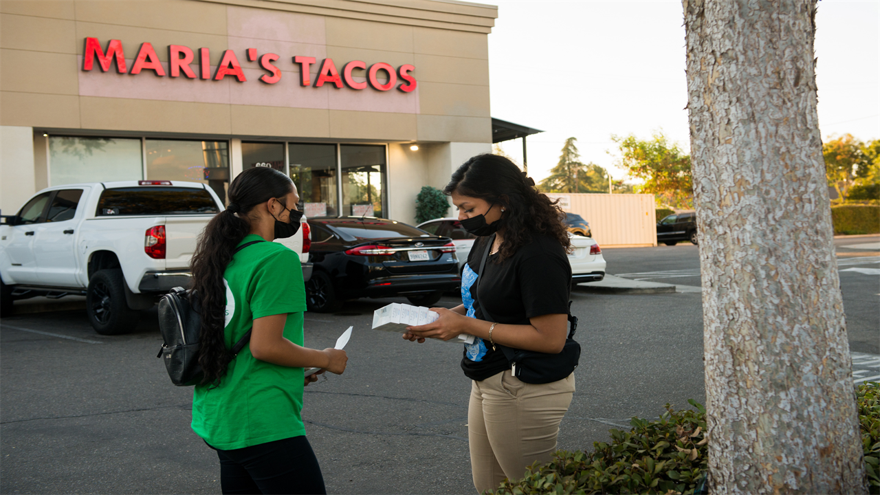 Melissa Lopez (left) and Alma Gallegos (right) get ready to distribute covid tests. They are standing in the parking lot in front of a building that says "Maria's Tacos."