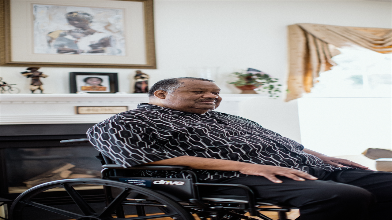 Thomas Greene is sitting in a wheelchair in his home, which is brightly lit by sunlight. He looks away from the camera.