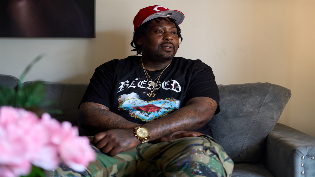 A man in a black t-shirt with the word 'Blessed' across the chest in white, looks to the left with a sombre expression on his face. His arms are tattooed and he has a gold watch around his wrist. 
