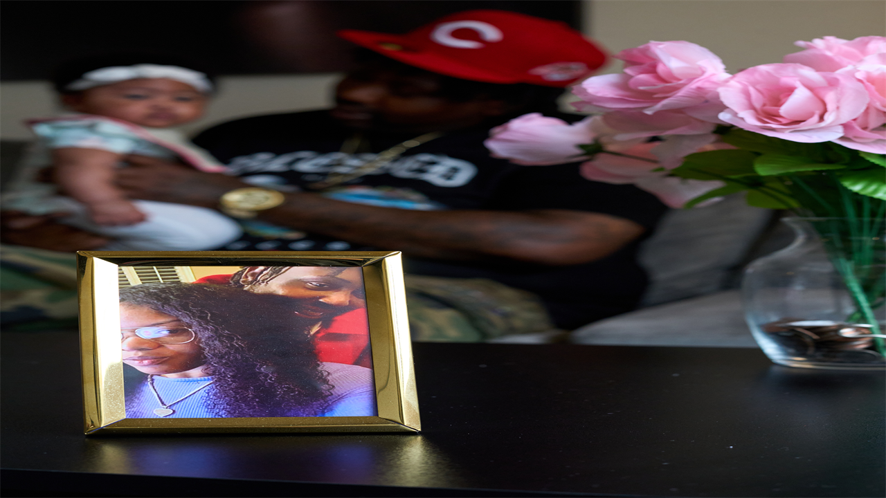 In the foreground is a vase of flowers and a gold framed photos of a man and woman on table. In the background, out of focus, is a father holding his baby daughter on his knee. 
