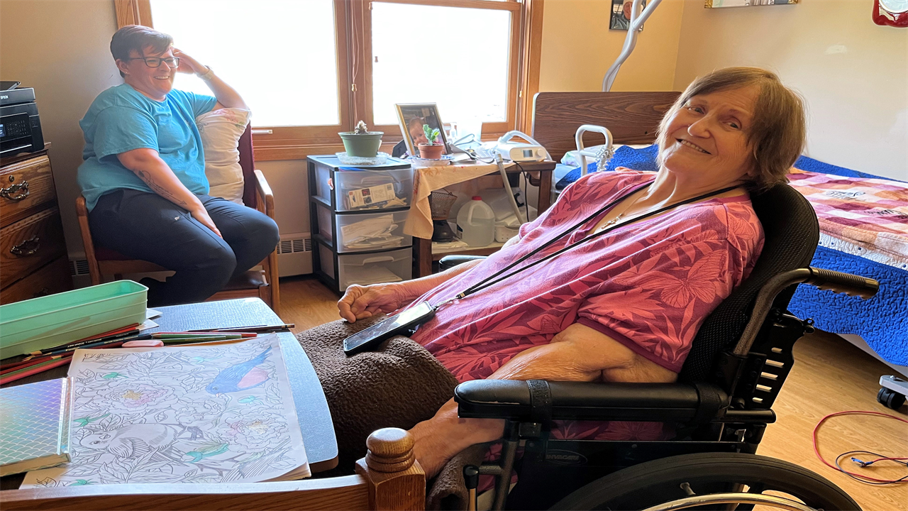 A photo of an older woman sitting at a desk in a wheelchair. Another woman is sitting in the background behind her.