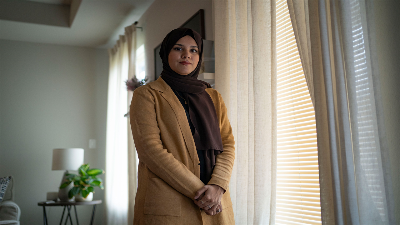 A woman stands next to a window and looks at the camera.