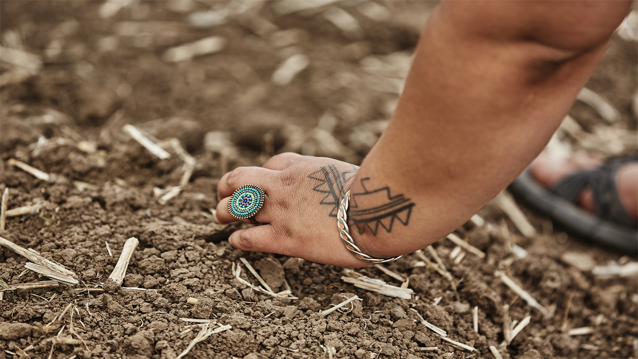 The tattooed hand of a woman, wearing a circular turquoise ring and a bracelet, digs in dirt in a garden.