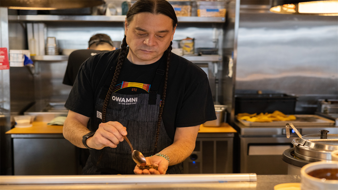 A man holds a spoon in his right hand over his cupped left hand as he stands in a restaurant kitchen wearing an apron