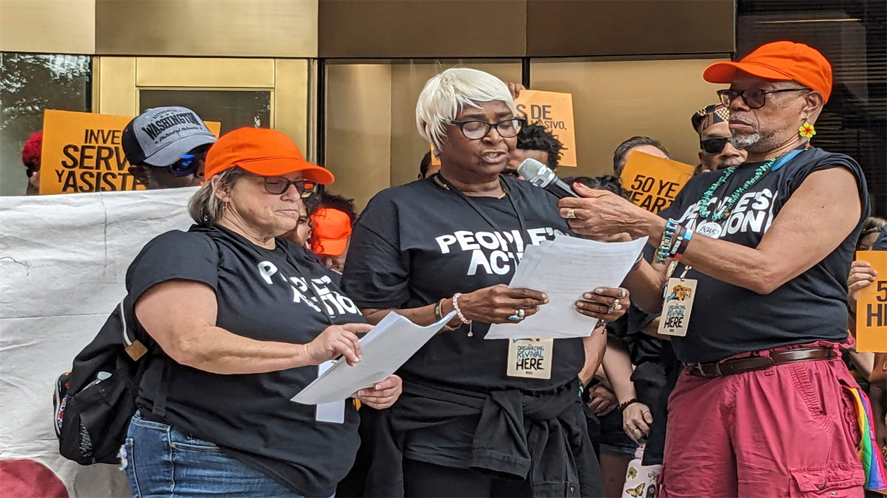A woman stands between two people and reads from piece of paper. The person to her left holds the microphone for her to speak into.