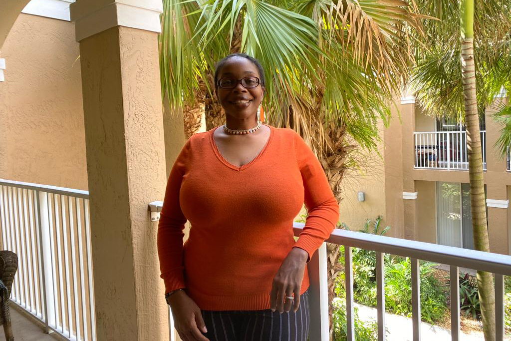 Phyllisa Deroze stand on an outdoor balcony in a warm climate. Palm trees are visible behind her as she smiles at the camera.
