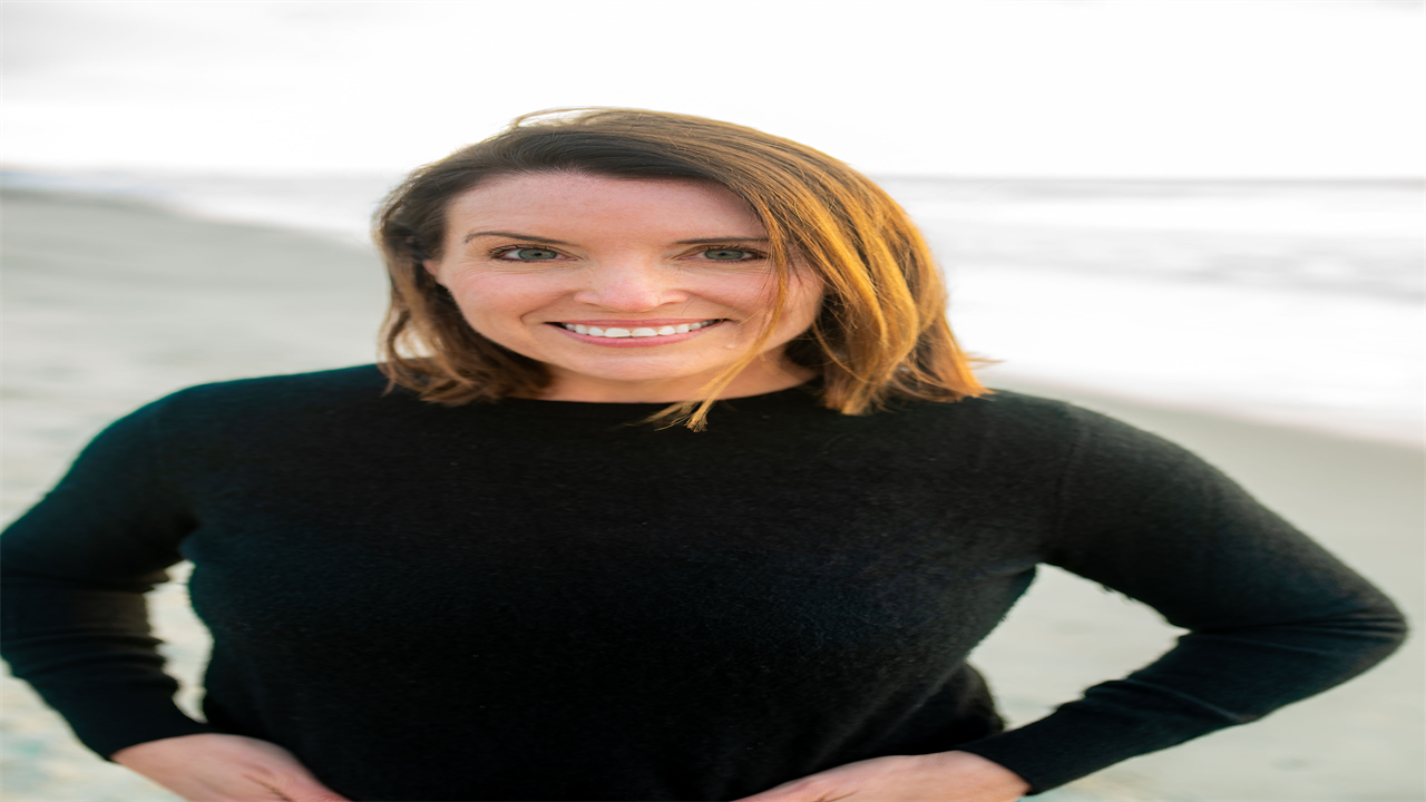 A portrait of Karla Adkins. She is standing outside on a beach and smiles at the camera.